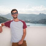 Young man in sunglasses leaning on a balcony with a scenic city view in the background.